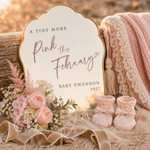 Könnte beinhalten: Ein weißes Acrylschild mit gewelltem Rand und Goldkante, das den Text A TINY MORE Pink this February BABY SWANSON 2027 trägt. Das Schild wird von einem Strauß aus rosa und weißen Blumen, einem Paar rosa Strickschühchen und einer Decke begleitet.
