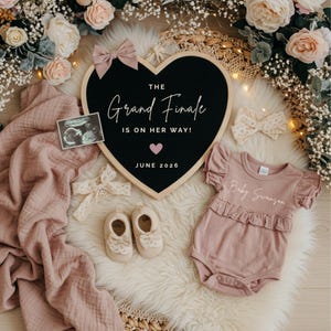 May include: A heart-shaped black felt letter board with the text THE Grand Finale IS ON HER WAY! JUNE 2026, a pink bow, a baby onesie with Baby Swanson, cream baby shoes, two lace headbands, a sonogram, and a dusty pink muslin swaddle blanket.