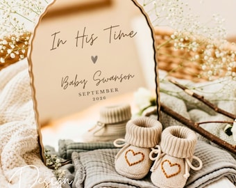 May include: A gold-edged mirror with the inscription "In His Time," a heart, "Baby Swanson," and "September 2026." Beside the mirror are two beige baby booties with embroidered hearts, resting on a gray blanket. The theme is baby announcement.