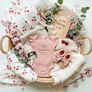 May include: A baby announcement basket with a pink onesie, baby shoes, blankets with cherry patterns, and a wooden sign that says "our little cherry on top". The onesie says "Baby Swanson August 2026".