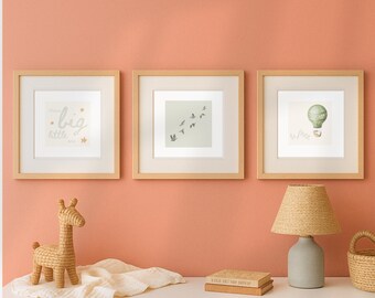 May include: Three framed prints with light wood frames hang on a peach wall. The prints feature a "dream big little one" text design, a flock of birds, and a hot air balloon. A woven giraffe, lamp, and books sit on a white surface.