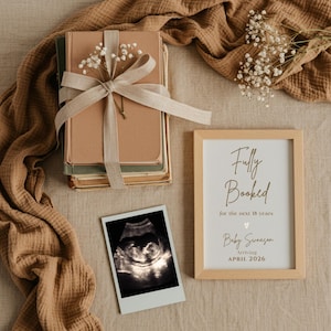 May include: A stack of books tied with a beige ribbon and decorated with white flowers. A framed sign reads "Fully Booked for the next 18 years" with a baby's name and arrival date. An ultrasound image is also present.