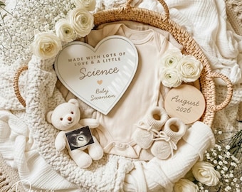 May include: A neutral-toned baby announcement arrangement in a woven basket. The arrangement includes a heart-shaped sign that reads "Made with lots of love & a little Science Baby Swanson", a teddy bear, baby booties, a onesie, and a wooden disc that reads "August 2026".