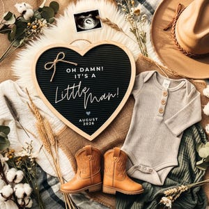 May include: A heart-shaped letter board with the text "OH DAMN! IT'S A LITTLE MAN!" and "AUGUST 2026". The image includes a baby's ultrasound, a tan cowboy hat, a gray onesie, and a pair of tan cowboy boots.