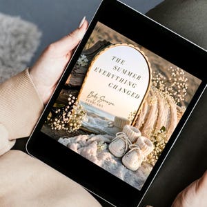 May include: A digital tablet displaying a graphic with an arched gold-rimmed mirror reflecting a beach scene. The text reads THE SUMMER EVERYTHING CHANGED, Baby Swanson, and FEBRUARY 2027. In the foreground are cream-colored knit baby booties.