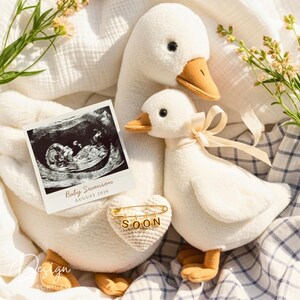 Puede incluir: Un par de juguetes de peluche de ganso blanco, uno grande y otro pequeño, con picos naranjas. Se muestra una foto de ultrasonido con el texto "Baby Swanson, August 2026". Se adjunta un alfiler de corazón de ganchillo con la palabra "SOON".