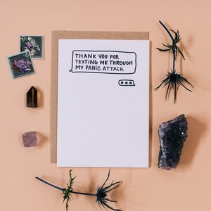 May include: A white card with black text that reads "Thank you for texting me through my panic attack." The card is surrounded by brown paper, a purple crystal, a brown crystal, and two sprigs of dried flowers.