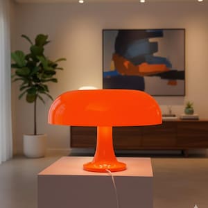 May include: A bright orange mushroom-shaped table lamp. The lamp features a rounded shade and a pedestal base. It sits on a white cube-shaped table. The lamp's cord is visible, and the background includes a plant, artwork, and a wooden cabinet.