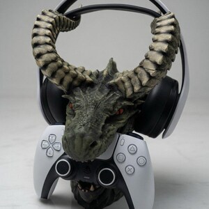 Può includere: Una scultura dettagliata della testa di un drago in tonalità verdi e grigie, che tiene in mano un controller di gioco bianco e nero. Un paio di cuffie bianche e nere poggiano sulle corna del drago. Un accessorio per il gaming.