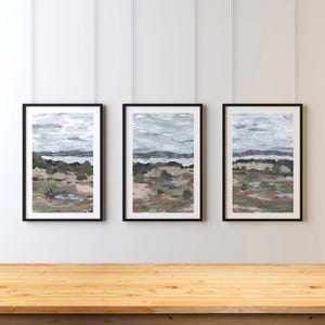 Może przedstawiać: Trzy oprawione obrazy krajobrazowe. Każdy z nich przedstawia teksturowane, abstrakcyjne przedstawienie naturalnej scenerii z wodą, lądem i niebem. Paleta kolorów obejmuje odcienie szarości, zieleni i stonowanej czerwieni.