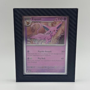 May include: A framed Pokémon trading card featuring Espeon, a purple creature with large ears. The card has text including "Psychic Assault" and "Psy Bolt". The frame is black with a carbon fiber pattern.