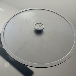 May include: Black round lid with a small black knob in the center. The knob has the word "000" embossed on it.