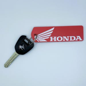 Op de afbeelding: Rode sleutelhanger met een wit Honda-logo en het woord "HONDA" in witte letters. De sleutelhanger is bevestigd aan een zwarte motorfiets sleutel.