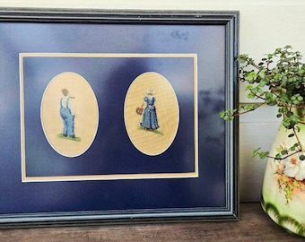 May include: Framed artwork featuring two oval cross-stitch portraits. One depicts a person in blue overalls, the other a person in a blue dress and bonnet. The frame is black with a navy blue matting.