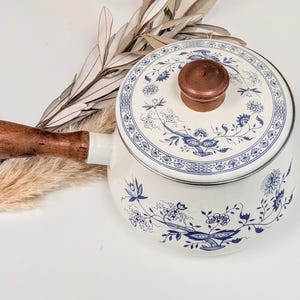 May include: A white enamel saucepan with a blue floral pattern, featuring a round lid with a brown wooden knob and a matching brown wooden handle. The pot is decorated with intricate blue botanical illustrations across the lid and body.