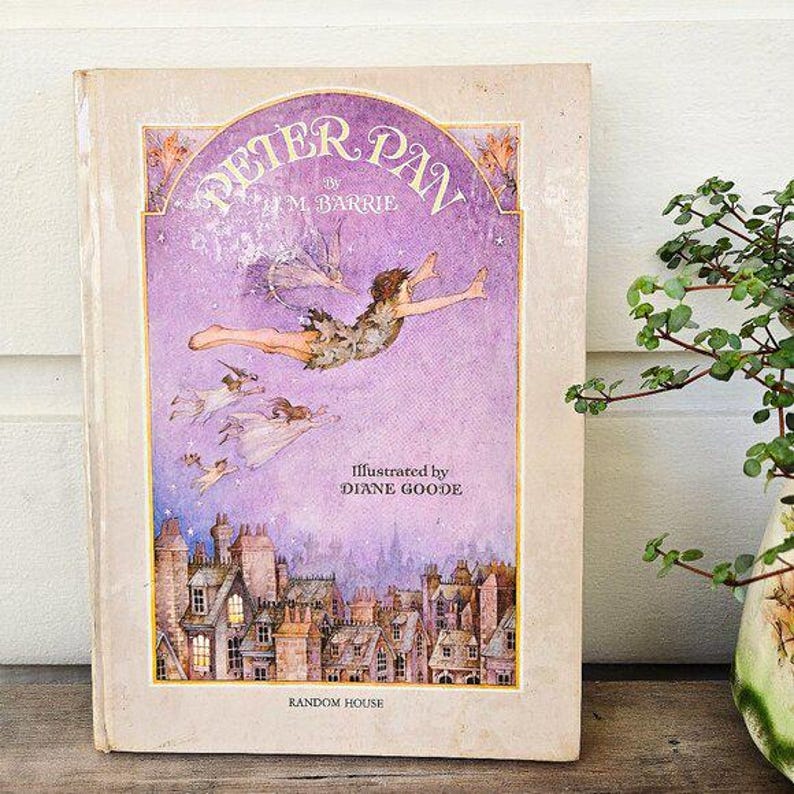 May include: A vintage hardcover book titled "Peter Pan" by J.M. Barrie, illustrated by Diane Goode. The cover features a purple background with an illustration of Peter Pan flying above a cityscape. The book is published by Random House.