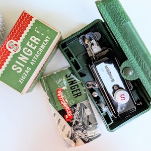 Peut inclure: Un accessoire de machine à coudre Singer pour boutonnières, noir, rangé dans un boîtier en plastique vert texturé, accompagné d'une boîte Singer pour zigzag rouge et verte et d'un manuel d'instructions Singer avec des illustrations.