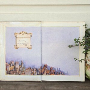 May include: Open book with a watercolor illustration of a townscape. The left page features a decorative frame with text, while the bottom of the page shows rooftops and a church steeple. The right page is blank. A vase with greenery is on the right.