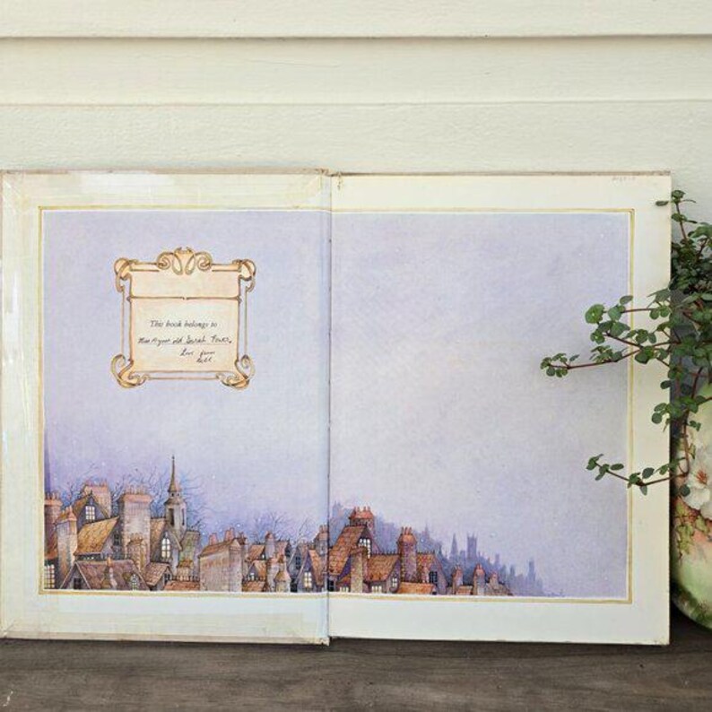 May include: Open book with a watercolor illustration of a townscape. The left page features a decorative frame with text, while the bottom of the page shows rooftops and a church steeple. The right page is blank. A vase with greenery is on the right.