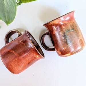 May include: Two ceramic mugs with a glossy, mottled finish in shades of deep copper, rust, and dark brown. Each mug features a curved handle and a slightly flared rim, showcasing a smooth, reflective surface with subtle variations in color tone.
