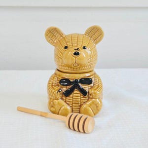 May include: A tan ceramic teddy bear-shaped honey pot with a black bow tie. It includes a matching lid and a wooden honey dipper. The bear has a textured design. The honey pot is on a white surface.