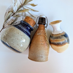 May include: Three ceramic vessels: a speckled beige and blue jar with a cork stopper, a tan bottle with a metal swing-top closure marked 13oz, and a small vase with a white neck and brown, blue, and tan horizontal bands on the body.