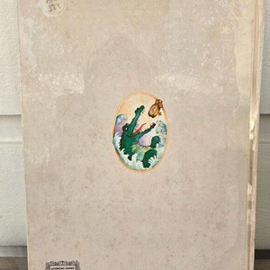 May include: A vintage book cover with a beige background and a colorful oval illustration. The illustration features a green crocodile emerging from blue and white waves, reaching for a small, golden clock. The book's title is not visible.