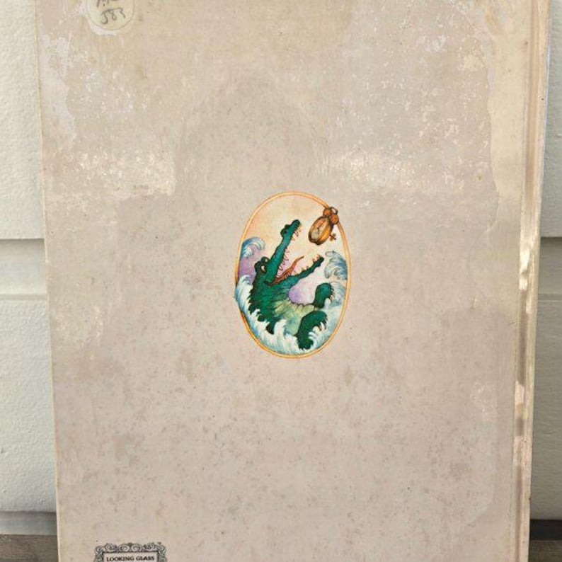 May include: A vintage book cover with a beige background and a colorful oval illustration. The illustration features a green crocodile emerging from blue and white waves, reaching for a small, golden clock. The book's title is not visible.