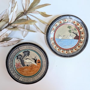Peut inclure: Deux assiettes rondes en céramique avec des motifs peints à la main. Une assiette présente un oiseau sur un fond orange texturé avec un arbre et une bordure géométrique. L'autre montre deux oiseaux sur une eau bleue avec un arbre et un bord floral.