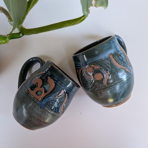 May include: Two rounded ceramic mugs featuring a dark blue and green glazed finish with exposed terracotta-coloured clay accents. Each mug has a curved handle and displays intricate, hand-carved patterns that reveal the natural clay beneath.