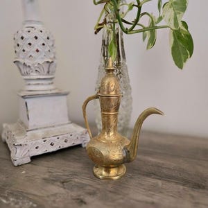 Könnte beinhalten: Eine kleine, verzierte Messingkanne mit Deckel und Ausguss. Die Kanne ist mit komplizierten floralen Mustern verziert, die in die Oberfläche eingeätzt sind. Die Kanne steht auf einem Holztisch.