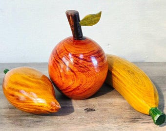 May include: Wooden fruit sculptures including an apple, mango, and banana. The apple is round with a brown stem and green leaf. The mango is teardrop-shaped, and the banana is curved. All are painted with realistic colors and wood grain.