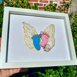 Puede incluir: Una obra de arte enmarcada con huellas de bebé azules y rosas, sobre un fondo de alas doradas y plumosas. La obra de arte está en un marco blanco, creando una pieza delicada y sentimental.