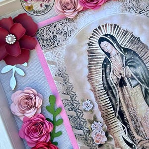Könnte beinhalten: Ein gerahmtes religiöses Kunstwerk mit einem Bild der Jungfrau Maria, umgeben von Papierrosen in Rosa- und Rottönen. Das Kunstwerk enthält dekorative florale Akzente und ein Logo in der oberen linken Ecke.