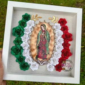 May include: A framed religious artwork featuring an image of the Virgin Mary. The frame is white, and the artwork is surrounded by paper roses in red, white, and green, resembling a flag. Gold leaf accents are also present.