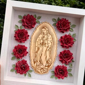 May include: A framed artwork featuring a wooden cutout of the Virgin Mary surrounded by eight red paper roses with green leaves. The artwork is set against a light gray background within a white frame.