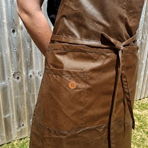 May include: A dark brown waxed apron featuring a smooth, water-resistant texture, a waist tie, and a single front pocket adorned with a small, circular, light brown leather patch embossed with a detailed star-like floral pattern design.