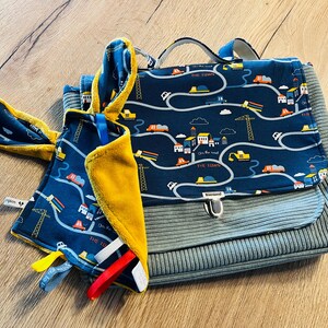 May include: A blue and yellow baby bag and matching blanket set. The bag features a flap closure and a handle. Both items have a repeating pattern of roads, cars, and construction vehicles. The blanket has colorful ribbons.