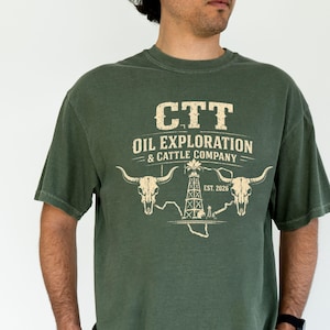 May include: Dark green t-shirt with a cream-coloured graphic. The design includes the text "CTT OIL EXPLORATION & CATTLE COMPANY", two bull skulls, an oil derrick, and the text "EST. 2026".