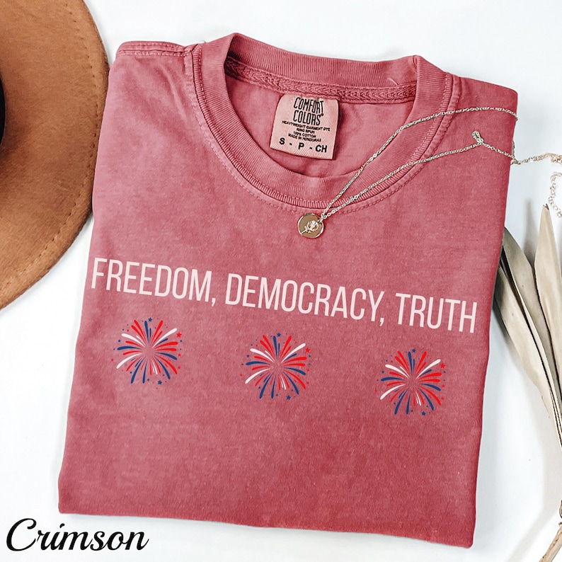 Pu&ograve; includere: T-shirt color cremisi con le parole bianche "FREEDOM, DEMOCRACY, TRUTH". La maglietta presenta tre disegni di fuochi d'artificio rossi, bianchi e blu. Una collana d'argento con un ciondolo &egrave; drappeggiata sulla maglietta.