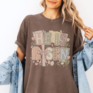 May include: Brown t-shirt with the text "HE IS RISEN" in a colorful, patchwork design. The design includes a cross, butterflies, flowers, and Easter eggs, perfect for Easter celebrations.