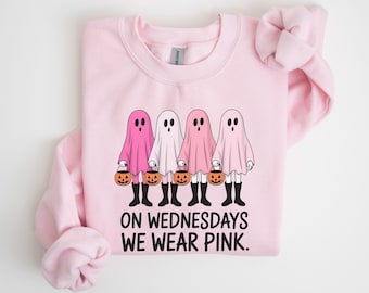 May include: Pink sweatshirt featuring four ghost figures holding jack-o'-lanterns. The ghosts are in shades of pink and white. The text "ON WEDNESDAYS WE WEAR PINK" is printed below the ghosts. The sweatshirt has a crew neck and long sleeves.