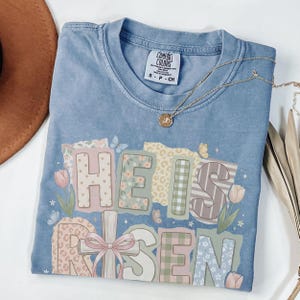 May include: A light blue Comfort Colors t-shirt with the words "HE IS RISEN" in a patchwork design. The design includes floral, plaid, and leopard print patterns. A gold necklace with a pendant is draped across the shirt.