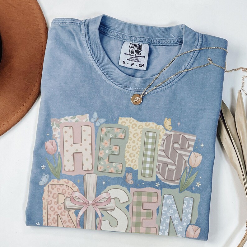 May include: A light blue Comfort Colors t-shirt with the words "HE IS RISEN" in a patchwork design. The design includes floral, plaid, and leopard print patterns. A gold necklace with a pendant is draped across the shirt.