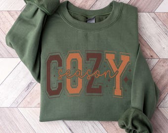 May include: An olive green sweatshirt with the words "COZY season" printed on the front in brown and orange. The sweatshirt has a crew neck and long sleeves with ribbed cuffs. The color is described as Military Green.