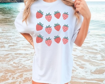 May include: White t-shirt featuring a pattern of red strawberries with green leaves. The strawberries are evenly spaced across the front of the shirt. The shirt is a casual, oversized style.