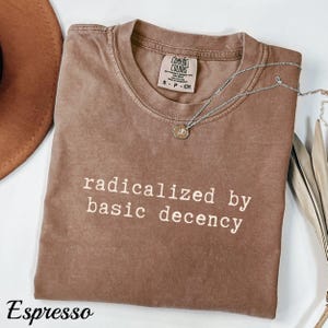 Pode incluir: T-shirt cor de café com a frase "radicalized by basic decency" em branco. Um colar de prata com um pequeno pingente repousa sobre a camisa. A camisa está dobrada e tem uma etiqueta Comfort Colors.