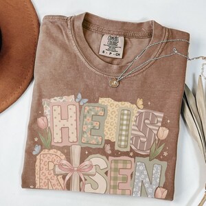 May include: Brown t-shirt with the words "He Is Risen" in a colorful, patchwork design. The design includes floral patterns, butterflies, and a cross with a pink bow. A silver necklace with a pendant is also visible.