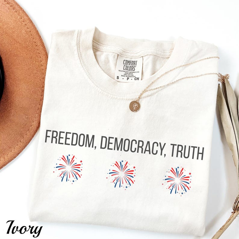 Pu&ograve; includere: T-shirt color avorio con le parole "FREEDOM, DEMOCRACY, TRUTH" stampate in nero. Sotto il testo ci sono tre disegni di fuochi d'artificio rossi, bianchi e blu. Sono visibili anche una collana d'oro e un cappello marrone.