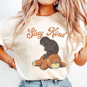 Pode incluir: Camisola creme com um gráfico de um macaco preto sentado em um macaco de pelúcia marrom. O texto "Stay Kind" está arqueado acima dos macacos em uma fonte marrom de estilo vintage. A camisola tem gola redonda e mangas compridas.